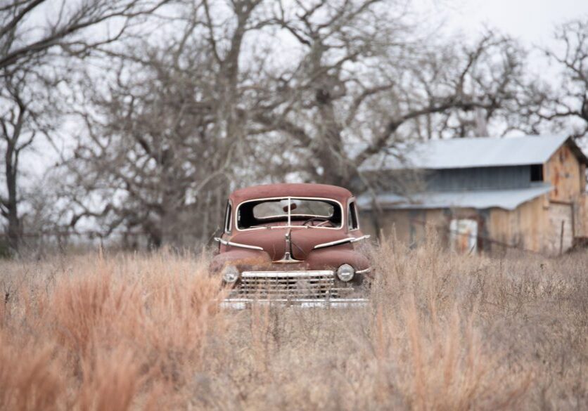 Rusty car parked in the middle of a field, surrounded by tall grass and trees